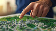 © pattaphorn - A hand points at a detailed architectural model, showcasing buildings surrounded by greenery, highlighting urban planning and design.