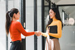© kenchiro168 - Two women in colorful sweaters shake hands in modern office space, showcasing professional interaction. Their smiles reflect positive and collaborative atmosphere