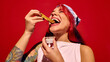 © master1305 - Young redhead woman in hat enjoying delicious churros dipped in hot chocolate sauce, set against bright red background. Concept of street food, winter fairs, pop art, holiday snacks