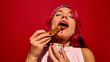 © master1305 - Young redhead woman passionately eating churros dipped into chocolate, set against bright red background. Concept of street food, winter fairs, pop art, holiday snacks
