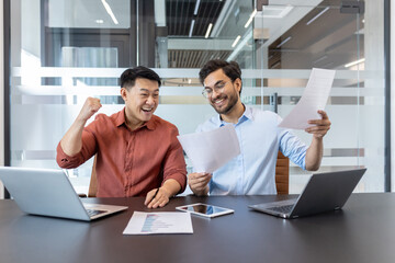 Wall Mural - Business colleagues celebrate success with laptops and documents. Smiling coworkers review project results on paper and phone, highlighting teamwork and achievement in modern office.