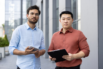 Poster - Two businessmen standing confidently outside modern office building, holding tablet and clipboard, ready for discussion. Their smart casual attire blends professionalism with style.