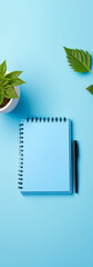 Aufkleber - A blue notepad with a pen and plant sit on an isolated pastel blue background leaving plenty of empty space around them.