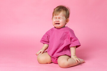 Wall Mural - Little child baby in a pink bodysuit sitting and crying isolated on a pink background. Childhood concept. High quality photo. Copy space