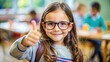 © Katewaree - Young girl smiling and giving a thumbs-up gesture in a classroom setting with classmates engaged in learning activities in the background, showcasing happiness and confidence.