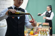 © DragonImages - Waiter pouring red wine into glass at elegant outdoor event with food and drinks. Background features another server preparing refreshments