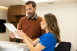 © Dejan - In an office setting, a man in brown sweater and woman in blue uniform study documents on a marble desk attentively, engaging in a professional discussion.