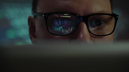 A person sits at a desk, focused on a computer screen displaying colorful data visuals. The dim lighting emphasizes the glow from the screen, creating a serious atmosphere