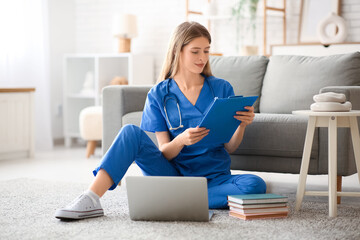 Wall Mural - Female medical intern sitting on floor with laptop and clipboard in living room