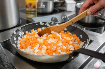 Wall Mural - A close-up of a frying pan on a stove, filled with diced butternut squash and onions being sautéed. A wooden spoon stirs the colorful mixture in a well-equipped kitchen.