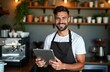 © miss irine - Cheerful barista uses tablet in cozy coffee shop. Man is smiling. He wears apron. Coffee shop interior is rustic and trendy. He is working at a counter. Image shows employee working in cafe.