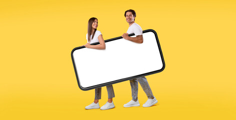 Poster - Cool Mobile App. Cheerful young couple holding big giant smartphone with mockup, walking together, promoting application or website, advertising your service or product over orange studio background