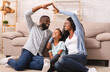 © Prostock-studio - Family Care And Protection Concept. Black parents making symbolic roof of hands above their little daughter, sitting on floor at home