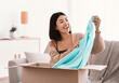 © Prostock-studio - Mail Delivery, Shipment And People Concept. Happy asian woman unpacking parcel, sitting on couch and holding clothing, unboxing cardboard package, copy space. Lady satisfied with purchase