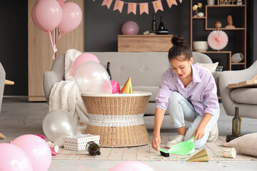 Young Asian woman sweeping confetti from carpet in messy room after Birthday party