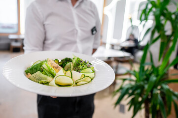 Wall Mural - A waiter presents a beautifully arranged plate of fresh salad with greens and cucumber ribbons. The setting is modern and elegant, perfect for showcasing culinary excellence.