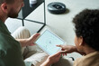 © pressmaster - Person sitting on sofa, holding and using a tablet, controlling smart home device in modern living room. Visible robotic vacuum cleaner on floor