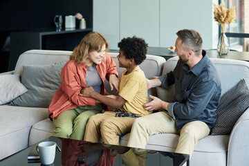 Wall Mural - Multiracial family sitting on a comfortable couch in a modern living room, sharing smiles and engaging in conversation, demonstrating warmth and connection