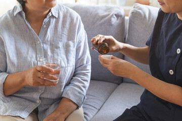 Asian young woman caregiver, carer support elderly, aged patient sit on sofa at home, holding glass of water with take medicine, cure or treatment disease of grandmother after retirement, help support