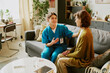 © AnnaStills - Healthcare worker sitting beside elderly lady in cozy living room providing comfort and care, indoor setting with natural light filtering through windows