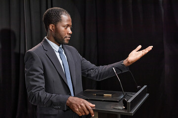 Medium shot of African American male politician in formal attire gesticulating with hands addressing audience while giving public speech on stage at podium against black curtains, copy space