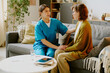 © AnnaStills - Caregiver in blue uniform sitting with senior woman wearing glasses, holding her hand, and offering emotional support in cozy living room with warm decor