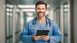 © mahmud - Cheerful handsome surgeon doctor man in blue uniform holding digital tablet computer, looking at camera, smiling, posing for portrait in clinic hall