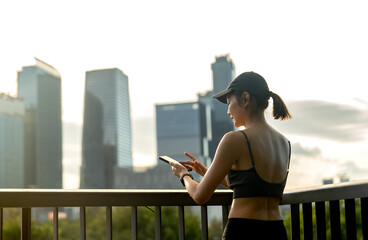 Wall Mural - Back of Asian sport woman use mobile phone and relax after exercise in public garden with high building of big city in background and soft light.