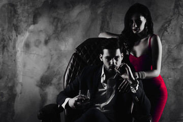 Poster - Portrait of brutal gentleman in formal wear sitting in leather chair holding glass with whiskey cigar in hands, charming pretty lady standing near looking at camera isolated on grey background
