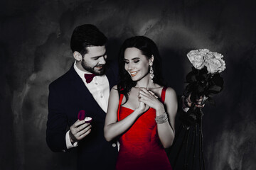 Aufkleber -  Portrait of stylish gentleman with stubble giving gold ring with diamond to excited cheerful lady with jewelry holding white roses in hands, couple isolated on grey background