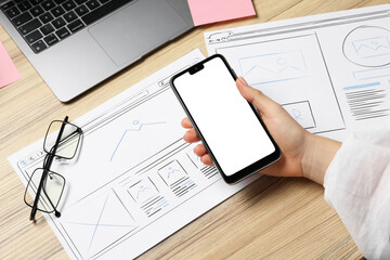 Leinwandbilder - Website designer holding smartphone at wooden table with wireframes and laptop, closeup