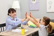 © New Africa - Autism therapy. Smiling psychologist giving high five to little girl at table in mental health center