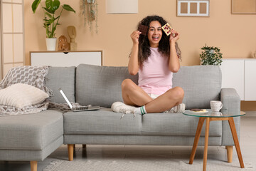 Poster - Happy young African-American woman with different bars of chocolate sitting on sofa at home