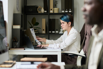 Poster - Young female trader with financial paper typing on laptop keypad while analyzing economical data by her workplace