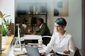 Poster - Young serious businesswoman with financial paper sitting by desk in front of laptop and scrolling through online information