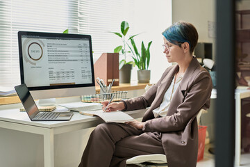 Aufkleber - Serious young woman in formalwear taking notes in copybook while sitting by workplace with desktop computer and laptop