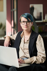 Aufkleber - Young elegant businesswoman in eyeglasses and formalwear looking at camera while sitting on sofa and using laptop in office