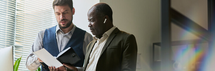 Poster - Banner of young male economist with open notepad explaining data on paper document held by mature African American colleague
