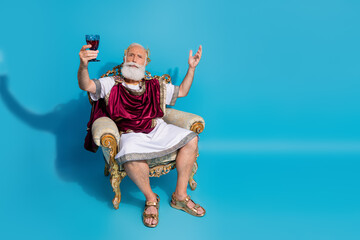 Full body portrait historical character costume man sit throne hold wine glass empty space of isolated on blue color background