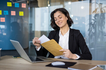 Wall Mural - Businesswoman enjoys moment opening envelope at desk, sitting with laptop in modern office. Scene conveys productivity, success, communication, professional environment