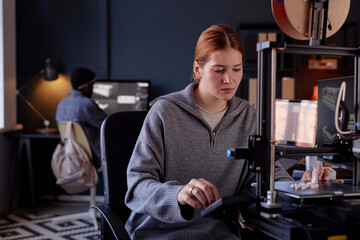 Fototapete - Woman sitting at desk working with 3D printer while colleague is in background focusing on his screen showing a busy and tech-savvy environment