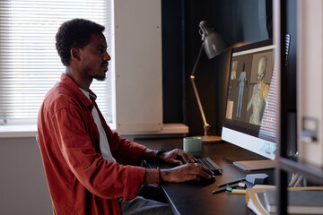Fototapete - Man sitting at desk, working on desktop computer in modern workspace, focusing on screen. Light entering from window at side, lamp on desk, and various items surrounding him