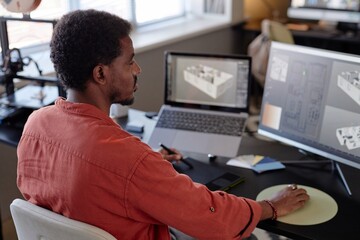 Fototapete - African American man focusing on creating architectural designs on large screen in modern office setting. Multiple digital devices and papers on desk supporting detailed work environment