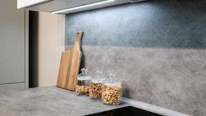 Poster - Countertop with wooden cutting board and three glass jars with nuts. Jars filled with mahogany peanuts in kitchen interior