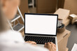 © fizkes - Small business owner woman using laptop with blank screen over stacked boxes, working in ecommerce Internet shop storage. Empty display of computer with copy space for logistic application interface