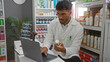 © Krakenimages.com - Handsome young man in a pharmacy examining a product and using a laptop in the store's interior workspace