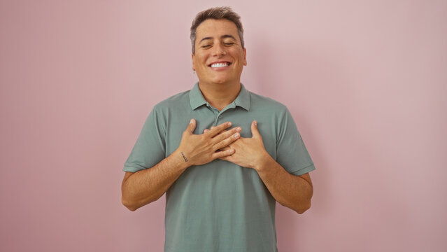 Handsome young man smiling with eyes closed and hands on heart, standing over an isolated pink background in a soft green shirt, embodying gratitude and joy in a candid portrait