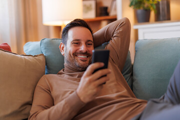 Wall Mural - Portrait of handsome mid adult man lying on sofa and texting online over smart phone in living room