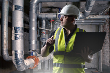 Poster - man engineer in basement. worker stands with laptop near heating pipes. dark basement with boiler pi