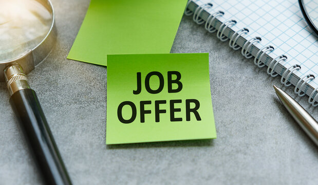 Job Offer text on green sticky note with magnifier, pen and notepad on gray office background.